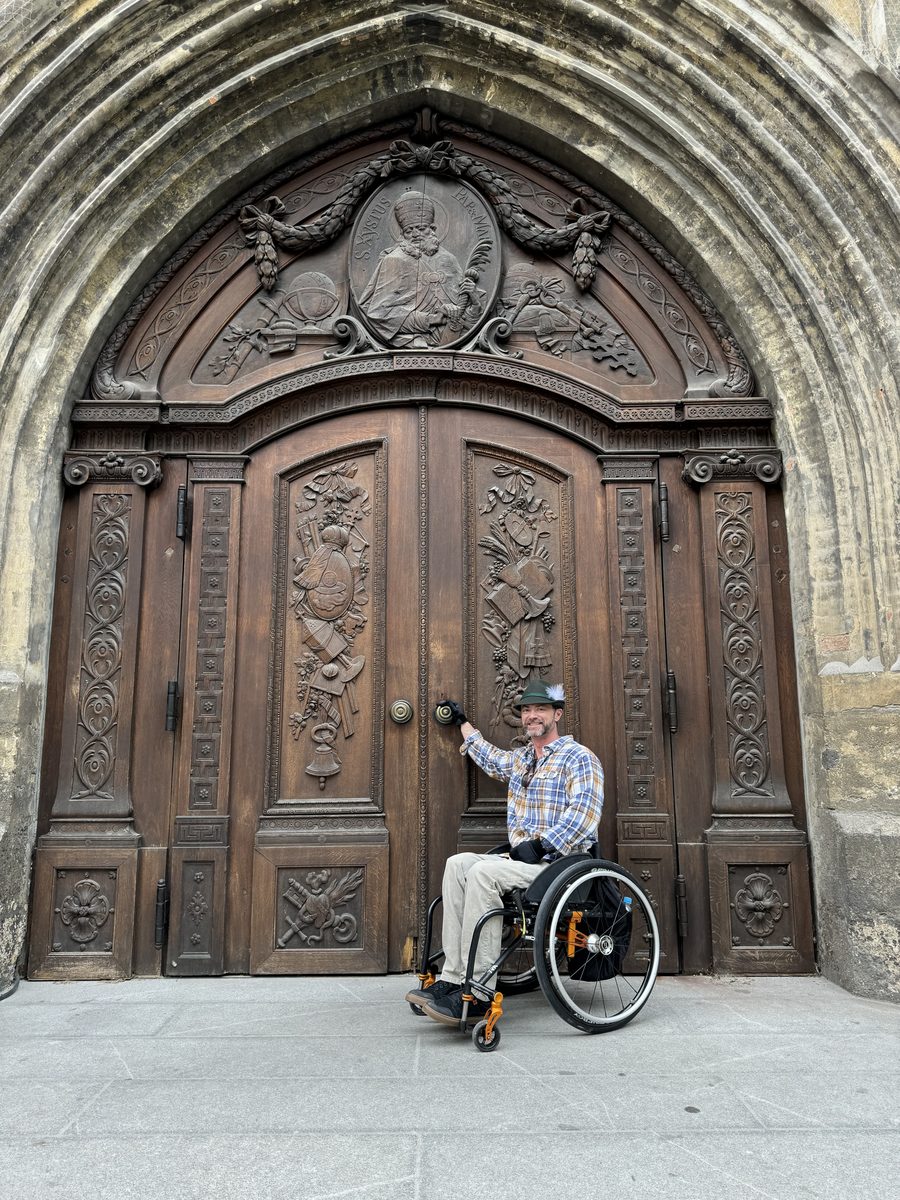 The host in his wheelchair, smiling in front of an ornately carved wooden cathedral door, tipping his hat.