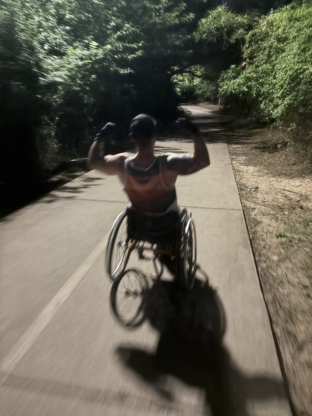 The host, a wheelchair user, raising both arms in triumph while wheeling down a tree-lined path.