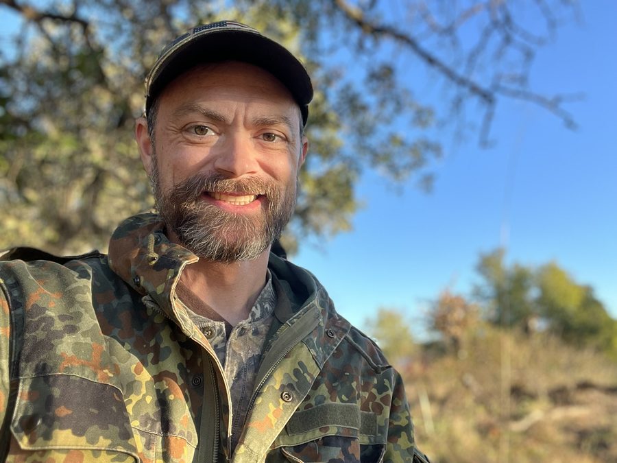The host smiling outdoors in a camouflage jacket and American-flag cap.